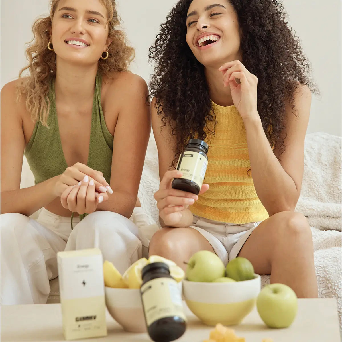 2 young women are eating GIMMY vitamin gummies while smiling. In front of them is a table with fresh fruit and the GIMMY Energy bottle and box on it.